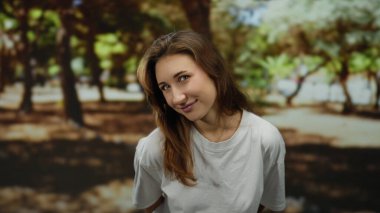 Güneşli parkta gülümseyen bir kadın ağaçlarla çevrili açık havada yaz atmosferini kucaklayan beyaz gömlekli doğal görünüşlü bir kadın günün tadını çıkarıyor..