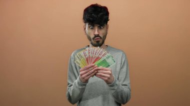 Elinde Norveç banknotları olan gri kazaklı bir adam kahverengi arka plana karşı heyecanlı bir şekilde izole edilmiş, yakışıklı bir ifadeyle para sergiliyor.