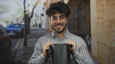 Güneşli bir sokakta siyah bir çanta tutan sakallı genç adam ağaçlar ve binalarla dolu neşeli bir açık hava sahnesi sergiliyor.