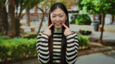 Çizgili tişörtlü bir kadın güneşli bir şehir sokağında arka planda ağaçlarla durup neşeli ifadesiyle mutluluğu ve pozitifliği ifade ediyor..