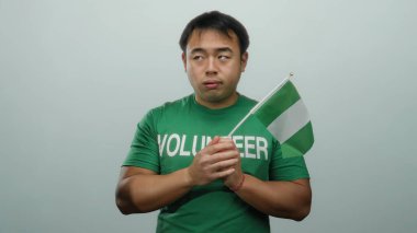 Beyaz arka planda Nijerya bayrağı taşıyan bir adam yeşil gönüllü tişörtü giyiyor ve ciddi bir ifade takınıyor..