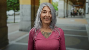 Pembe kazaklı, gri saçlı ve dövmeli yaşlı bir kadın. Açık bir şehir caddesinde gülümsüyor. Kemer ve bulanık şehir geçmişi olan, kendine güveni ve tarzı olan.