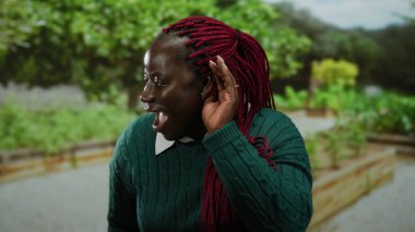 Parkta yeşil süveter giyen Afro-Amerikalı bir kadın, eli kulağında, yemyeşil bir zemin ve sakin bir arka planla çevrili, dikkatle dinlemeyi öneriyor..