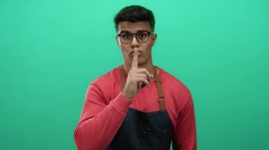 Gözlüklü ve kırmızı önlüklü genç adam, yeşil bir arka plana karşı sessiz, kavramsal iletişim için izole edilmiş, ciddi bir ifadeye sahip..