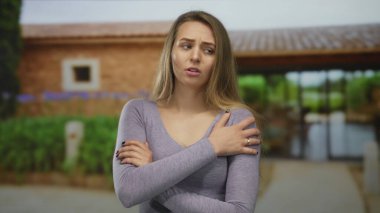 Dışarıda genç bir kadın, yeşil ve mavi çiçeklerle çevrili kırsal bir binanın önünde kolları çaprazlanmış sıradan bir kıyafet giyiyor..