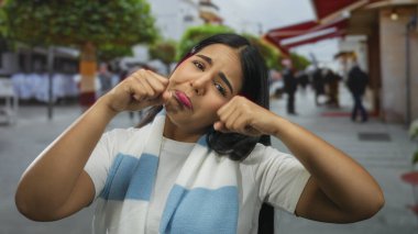 Sokakta üzgün bir ifade sergileyen kadın, atkı ve tişört giyiyor, çevresi kentsel çevre ve ağaçlarla çevrili, canlı bir ortamda duygularını iletiyor..
