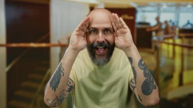 Sakallı adam, bir otel lobisinde şakacı bir şekilde yüzünü örten, karizma sergileyen ve kapalı mekanda çekici bir kişiliğe sahip..