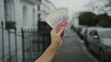 Şehir sokaklarında Arjantin pesoları tutan adam, canlı banknotları bulanık bir şehir geçmişine karşı sergiliyor, para birimini ve ekonomik temaları vurguluyor..