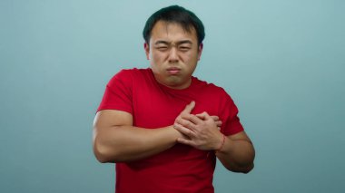 Kırmızı gömlekli adam göğsünü mavi arka planda sıkıntılı bir şekilde tutar. Gergin bir ifadeyle bir rahatsızlık veya endişe anını tasvir eder..