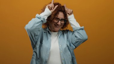 Kadın neşeli boynuzlu jest yapıyor, parmakları turuncu arka planda, neşeli ve eğlenceli bir ifade takınıyor, günlük kot gömlek ve gözlük takıyor..