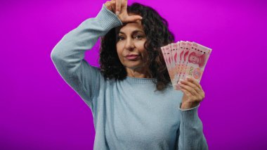 Bir kadın Çin Yuan banknotlarını tutarken ezik jestler yapıyor, izole edilmiş mor duvarlara karşı duruyor, duygularını ve finansal kavramlarını ifade ediyor..