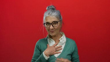 Gri saçlı ve yeşil gözlüklü yaşlı kadın canlı kırmızı bir arkaplanda poz veriyor, elleri göğsünde şükranlarını sunuyor, sıcaklık ve güven saçıyor..
