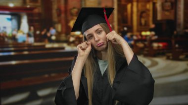 Mezuniyet kıyafetli bir kadın, arka planda bulanık iç kısımları olan büyük bir kilisenin içinde hüzün ile alay ederek mizahi bir jesti vurguluyor..