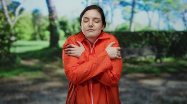 Turuncu hapishane tulumlu esmer kadın yemyeşil bir park ortamında huzur içinde kendini kucaklıyor, iç gözlem ve yalnızlık yapıyor..