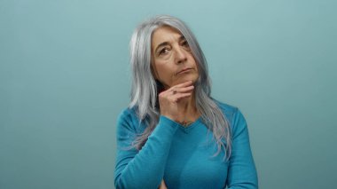 Gri saçlı yaşlı kadın mavi arka plana karşı jest yapıyor. Çevreden izole edilmiş düşünceli ve anlamlı bir şekilde durup düşünüyor.
