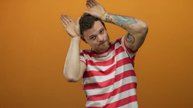 Çizgili tişörtlü genç İspanyol, izole edilmiş turuncu arka plana karşı neşeli kulak hareketi yapıyor..