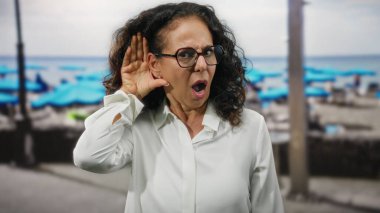 Deniz kenarındaki kıvırcık saçlı İspanyol kadın dikkatle dinliyor, şaşırıyor, plajda gözlük takıyor, merak ve ifadeyi vurguluyor..
