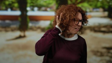 Parkta bir kadın, merak ve şaşkınlık ifadesiyle dinlerken orta yaşlı esmer İspanyol bir kadınla günlük giysiler içinde bir anlık etkileşim kuruyor..