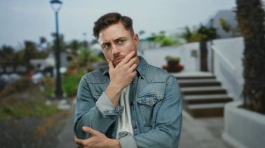 Sakallı genç bir adam, şehir dışında bir sokakta kot ceket giyerek derin derin düşünüyor..
