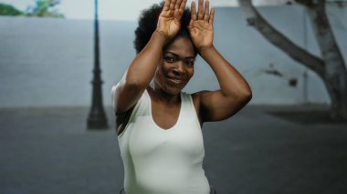 Afro-Amerikalı bir kadın, şehir sokaklarında elleri açık, beyaz bir atlet giymiş, bulanık şehir manzarası ve güneş ışığıyla çevrili, şakacı bir şekilde kulak hareketi yapıyor..