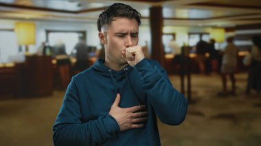 Mavi kapüşonlu genç adam arka planda yürüyen insanlarla dolu yoğun bir otel lobisinde öksürüyor..