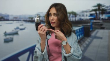 Elinde matkap tutan bir kadın, sahil kenarında yürürken gülümseyerek matkabı işaret ediyor..