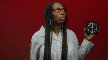 Gözlük ve çizgili tişört giyen, kırmızı arkaplanda saati tutan kadın yakın plan çekimde zaman kavramını aktarıyor.