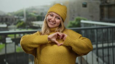 Roma harabelerinde sarı şapka ve süveter içinde gülümseyen kadın, kalp elleri yapıyor, şehirdeki antik taş arka planda yumuşak bir odak ile.
