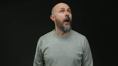 Sakallı bir adam, izole edilmiş siyah bir arkaplana şaşkınlıkla bakıyor gri bir süveter giyiyor. Hislerini minimalist ortamda ifade ediyor..