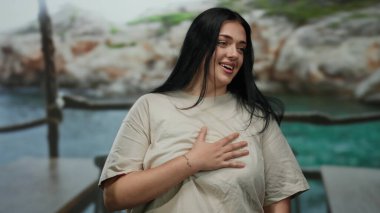 Sahilde gülümseyen kıvrımlı genç bir kadın. Arka planda kayalık kayalıklar var..