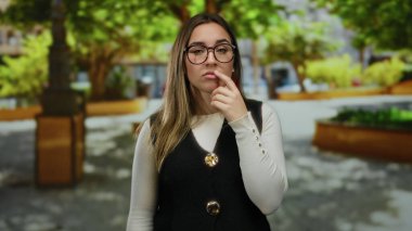 Gözlüklü genç bir kadın, dışarıda güneşli bir parkta yeşil ve ağaçlarla çevrili bir yelek ve beyaz gömlek giyerek düşünceli bir poz veriyor..