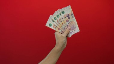 Arjantin pezosu tutan adam, izole edilmiş kırmızı arka planda beyaz eli zenginliği sembolize eden parayla gösteriyor..