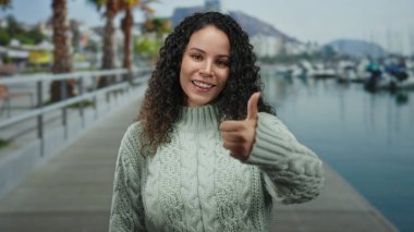Gülümseyen kadın, arka planda sakin bir deniz ve kayıklar olan palmiye ağaçlarının çerçevelediği manzaralı bir sahil gezisinde baş parmağını kaldırıyor..