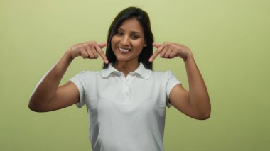 Genç İspanyol kadın, izole edilmiş sarı arka planı işaret ediyor. Beyaz tişörtlü, mutlu bir ifadeyle gülümsüyor..