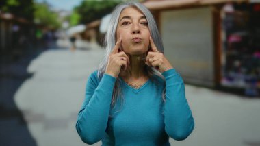 Gri saçlı, mavi kazaklı son sınıf öğrencisi bir kadın açık hava pazarında duruyor ve parlak bir sokak ortamında arka planda neşeli bir yüz ifadesi sergiliyor..
