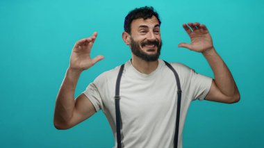 Sakallı İspanyol bir adam, pantolon askısı takıyor, neşeli ve neşeli bir enerji saçan, parlak mavi bir arka plana karşı şakacı hareketler yapıyor..