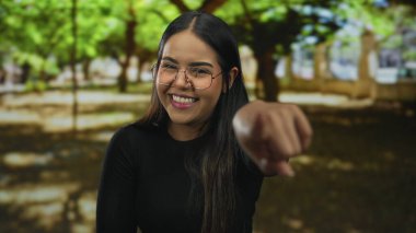 Sonbahar ağaçlarıyla parkta gülen, gözlüklü, siyah gömlekli, açık havada güneşli bir günde doğada neşeli bir ifade sergileyen bir kadın..