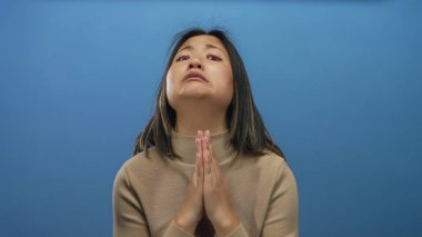 Elleri bağlı dua eden bir kadın mavi bir arkaplan karşısında umudunu dile getiriyor. İçten ve duygusal bir duruş sergiliyor..