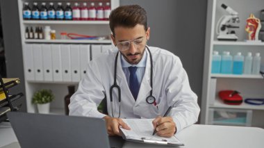 Genç İspanyol erkek doktor, arka planda raflar ve tıbbi ekipmanlarla bir hastane odasında notlar yazıyor.
