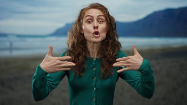 Sahilde enerjik bir şekilde el kol hareketi yapan, arka planda dağlar olan bir kadın, açık deniz kenarında iletişim ve ifadeye vurgu yapıyor..
