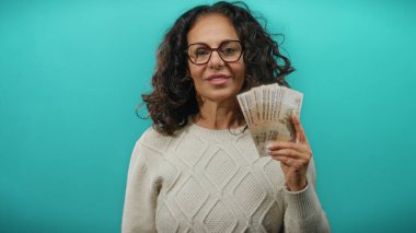 Orta yaşlı bir kadın elinde Rus rublesi banknotları tutuyor, mavi arka planda gözlük ve süveter takıyor, finans ve para birimi temalarını sergiliyor..