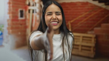 İç mekan inşaatında gülümseyen ve elini uzatan genç İspanyol kadın tuğlalar ve inşaat elementleri arasında samimiyet ve dostluk sergiliyor..