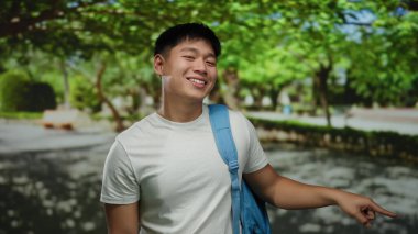 Serin yeşil bir parkta sırt çantasıyla gezen ve gülümseyen genç adam, neşeli ve neşeli bir eğlence ve keşif atmosferi sergiliyor..