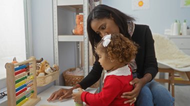 Evde kızını kucaklayan bir kadın samimi bir odada oyuncaklarla sevgi ve bakım gösteriyor aile bağlarını ve çevreyi yansıtıyor..