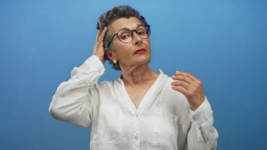 Gri saçlı, gözlüklü, beyaz bluz giyen bir kadın mavi arka planda poz veriyor, zarif ve kendine güvenen bir tavırla tarzını sergiliyor..