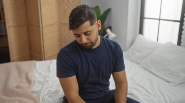 Genç adam rahat bir yatak odasında, düşünceli bir şekilde yatağında oturuyor, sıcak, davetkar bir iç mekan içinde bir içgözlem ve sükunet hissi aktarıyor, gündelik ama yakışıklı görünümünü vurguluyor..