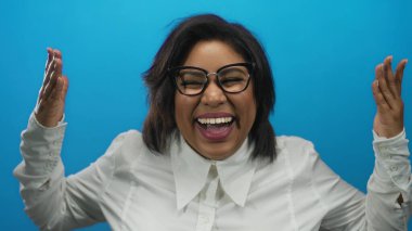 Gözleri kapalı gülen ve sonra kollarını kaldıran, mavi arka planda gözlük ve beyaz gömlek giyen, neşeli ruh hali gösteren bir kadın..