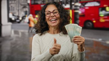 Başparmakları havada gülümseyen bir kadın, arka planda ikonik kırmızı otobüsler olan hareketli bir caddede, hareketli bir şehir ortamını yakalayan kirli para banknotlarını tutuyor..