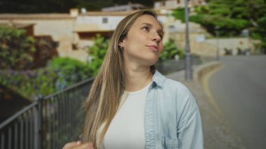 Güneşli bir günde, açık havada, uzun sarı saçlı, rahat gömlekli, yeşil ve kırsal mimariyle çevrili, rahat ve mutlu görünen bir kadın..