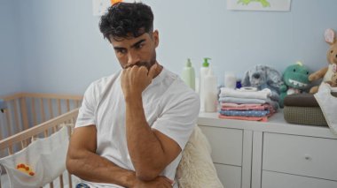 Sakallı genç bir adam beşikteki rahat bir yatak odasında dikkatlice oturuyor, bebek kıyafetleri ve oyuncaklarıyla çevrili, ev ortamını tasvir ediyor..